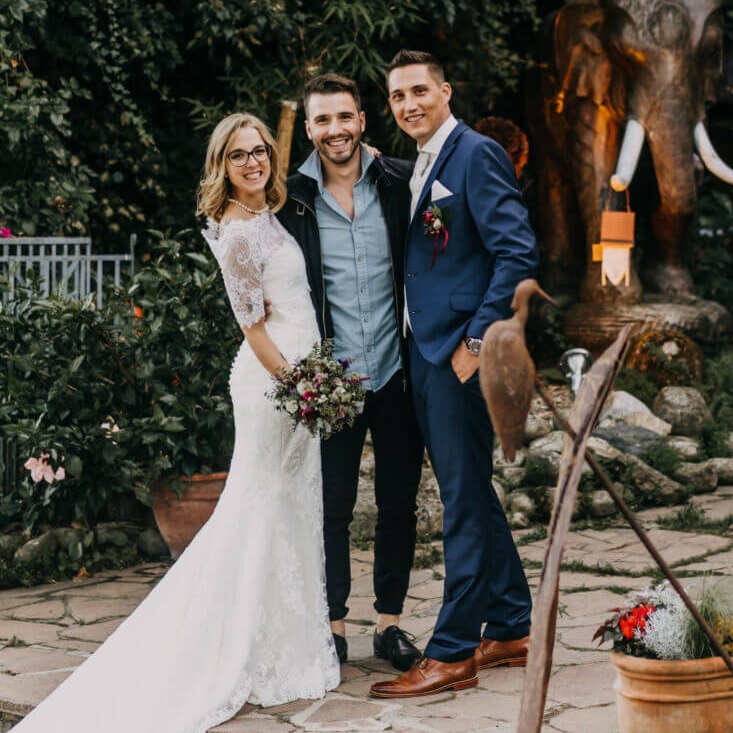 Eine Braut im weißen Kleid und ein Bräutigam im blauen Anzug stehen draußen auf Steinstufen. Sie werden von einem Mann in Freizeitkleidung flankiert, alle lächeln. Hinter ihnen ist üppiges Grün und eine steinerne Elefantenstatue mit weißen Stoßzähnen.