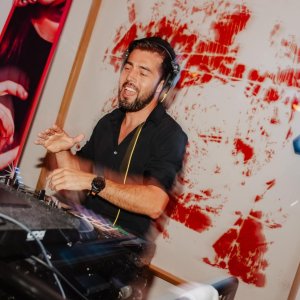 Ein DJ mit Kopfhörern lächelt und hebt eine Hand, während er in einem DJ-Pult Musik auflegt. Hinter ihm sind abstrakte rote Kunstwerke und ein Plakat mit "JET" und Veranstaltungshinweisen zu sehen. Die Szene sieht energiegeladen und lebendig aus.