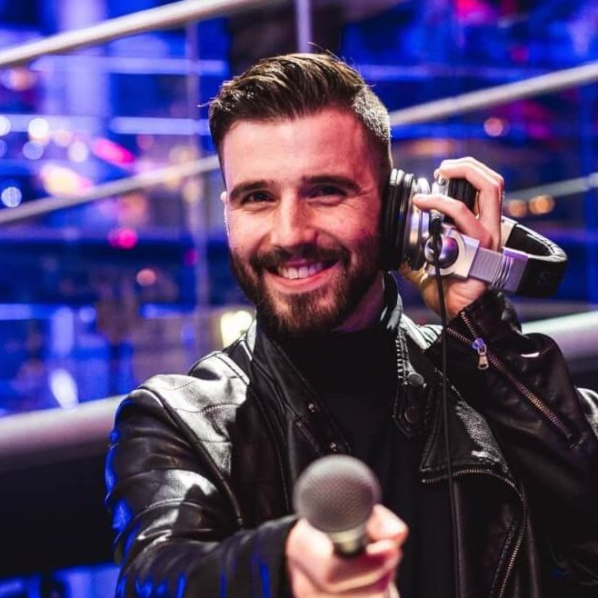 Ein Hochzeits- und Event-DJ steht in einer schwarzen Lederjacke mit Mikrofon und Kopfhörern hinter dem DJ-Mischpult. Vor dem Hintergrund bunter Lichter in der modernen, clubähnlichen Umgebung lächelt er begeistert.