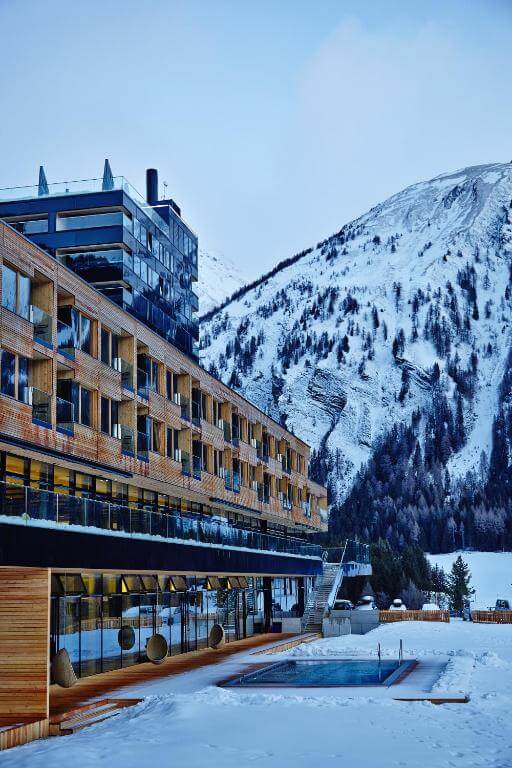 Ein modernes Hotel mit einer Kombination aus Holz- und Glasfassade liegt am Fuße schneebedeckter Berge. Im Vordergrund befindet sich ein Außenpool und der Boden ist mit Schnee bedeckt. Der Himmel ist klar und sorgt für eine ruhige Winteratmosphäre.