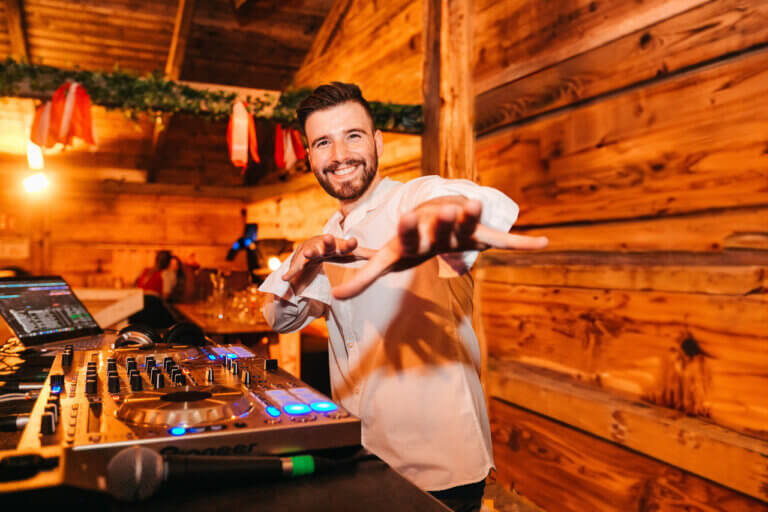 Ein lächelnder DJ im weißen Hemd tritt bei einer lebhaften Veranstaltung in einem warm beleuchteten Holzsaal auf. Er unterhält sich begeistert mit der Menge, streckt die Hände aus und steht hinter einem DJ-Pult mit Plattenspielern und einem Laptop. Von der Decke hängen Dekorationen.