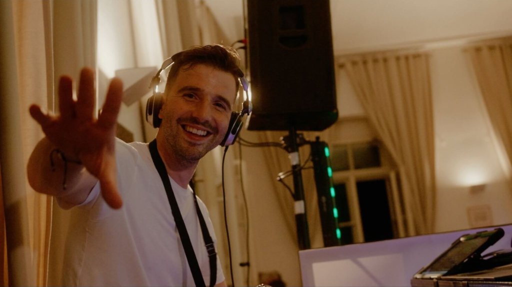 Hochzeits DJ Joseph Erin mit Kopfhörern und weißem Hemd lächelt und streckt die Hand in die Kamera. Sie befindet sich in einem Raum mit Vorhängen und steht neben einer Audioanlage und einem Lautsprecher.