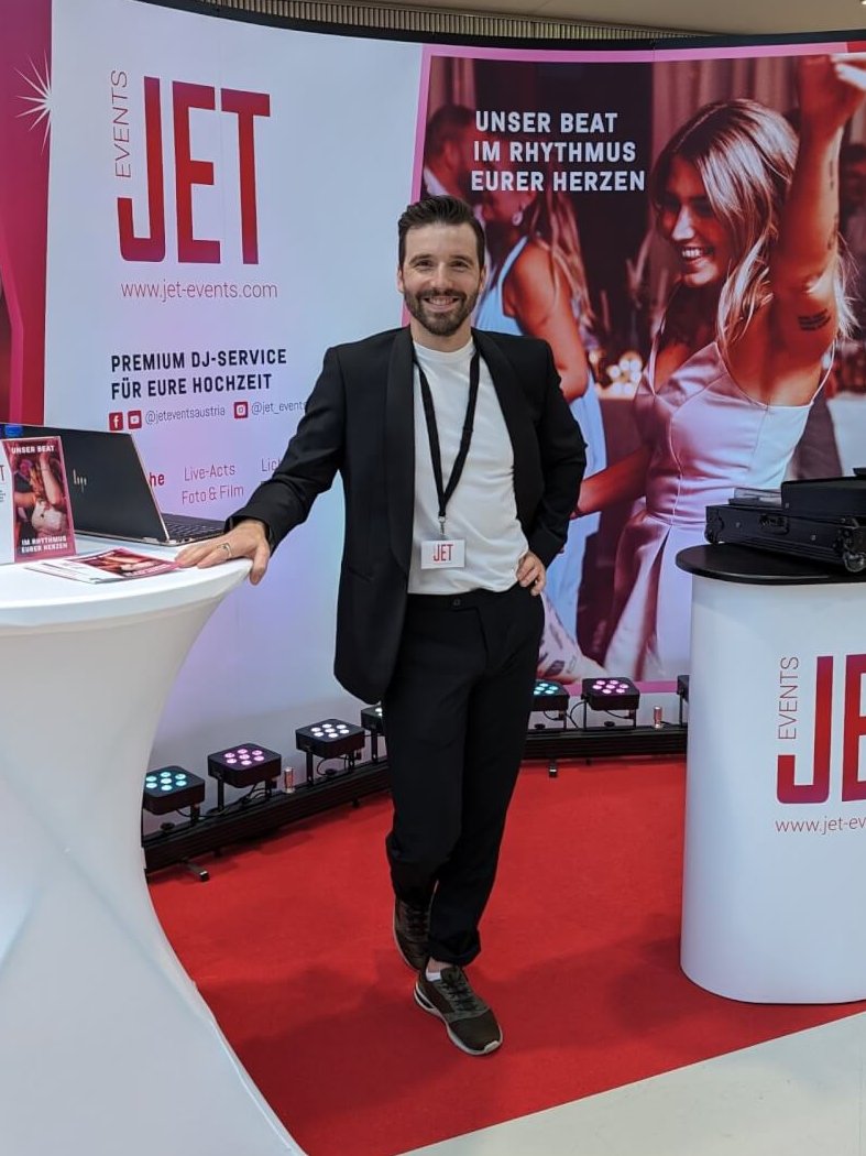 Ein Mann im Anzug steht lächelnd an einem Werbestand von JET Events. Hinter ihm hängt ein Banner mit einer tanzenden Frau und einem Text. Am Stand steht ein Tisch mit Werbematerialien und auf einer Theke ein Laptop.