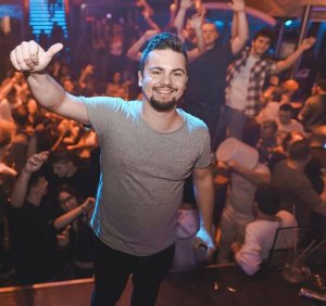 Ein junger Mann steht lächelnd in einem grauen T-Shirt in einem überfüllten Nachtclub und streckt den Daumen hoch. Um ihn herum tanzen die Leute und heben ihre Hände inmitten der blauen und orangeroten Lichter. Die Atmosphäre ist lebhaft.