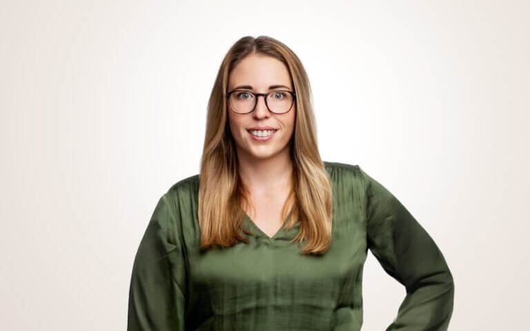 Eine Frau mit langen blonden Haaren, die eine Brille und eine grüne Bluse trägt, lächelt in die Kamera vor einem schlichten hellen Hintergrund.