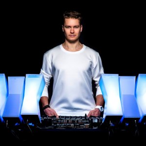 Ein Mann in einem weißen Hemd steht hinter einem DJ-Mischpult, umgeben von geometrischen, blau beleuchteten Tafeln, vor einem dunklen Hintergrund.
