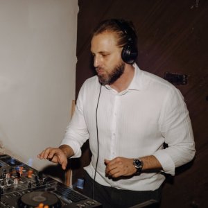 Ein DJ mit Kopfhörern und weißem Hemd bedient ein Pioneer DJ-Pult in einem schwach beleuchteten Raum. Er steuert die Geräte und konzentriert sich dabei auf die Musik. Im Hintergrund sind eine Ziegelwand und eine Holztür zu sehen.