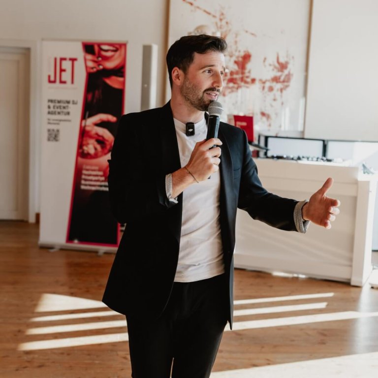 Ein Mann im schwarzen Anzug hält ein Mikrofon und gestikuliert, während er in einem hellen Raum mit Holzböden, moderner Kunst an den Wänden und einer stilvollen Atmosphäre spricht, die perfekt für Musik und DJs für Feiern in Österreich ist.