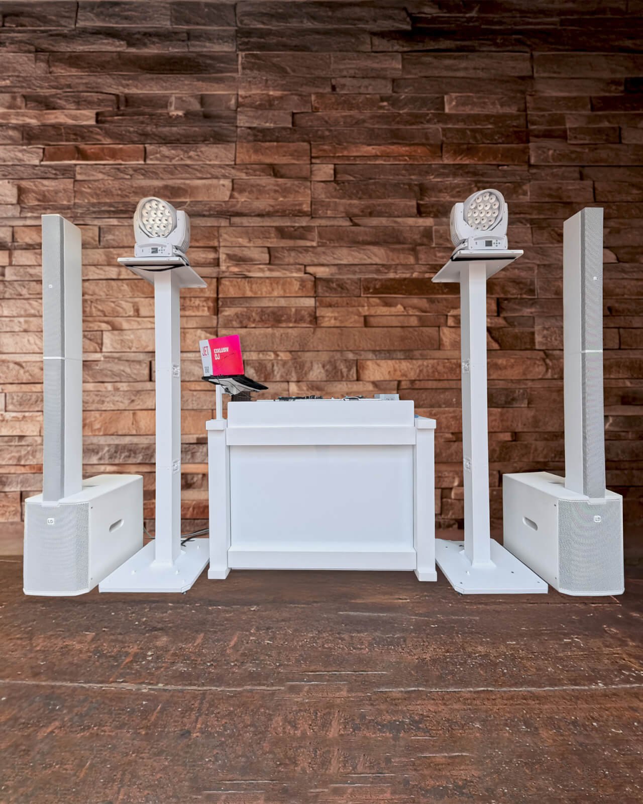 Ein modernes, weißes DJ-Pult mit Lautsprechern, Licht und DJ-Equipment steht auf einem Holzboden vor einem braunen Steinmauer-Hintergrund - perfekt für einen Hochzeits-DJ, der für die richtige Stimmung auf jeder Hochzeitsfeier sorgt.