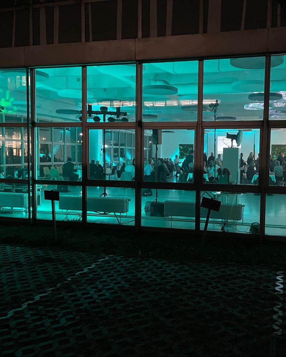 Große Fenster geben den Blick frei auf eine hell erleuchtete Indoor-Veranstaltung mit vielen versammelten Menschen, beleuchtet von türkisfarbenen Lichtern. Ein geschmückter Weihnachtsbaum steht in der Nähe der rechten Seite, perfekte Inspiration, wenn Sie eine Weihnachtsfeier planen wollen oder einen DJ für Firmen Weihnachtsfeier Österreich brauchen.