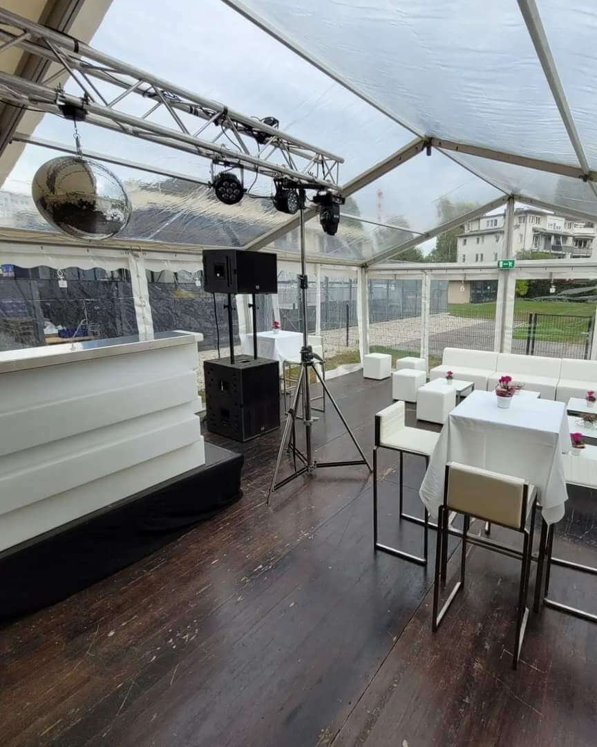 Ein modernes Outdoor-Event-Setup unter einem transparenten Zelt, perfekt für eine DJ-Firmen-Weihnachtsfeier Österreich, mit weißen Sofas, Stehtischen mit Hockern, einem Bartresen, Lautsprechern, Lichtequipment und einer Discokugel auf dunklem Holzboden mit Blick auf Gebäude und Grünflächen.