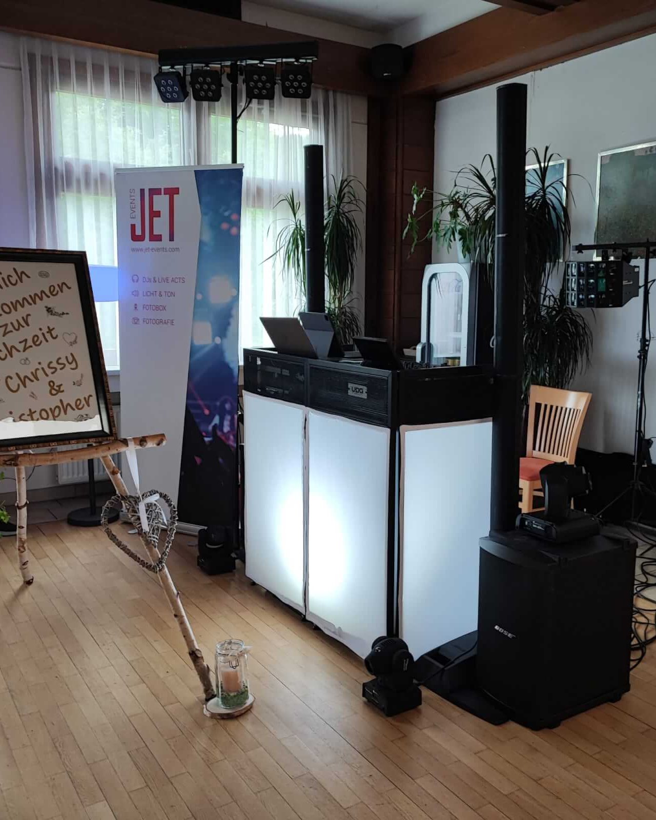 Ein DJ-Pult mit Lautsprechern, Lichtanlage und einem Holzstuhl ist drinnen aufgebaut - perfekt für eine DJ Firmen Weihnachtsfeier Österreich. Ein Willkommensschild und ein "JET"-Banner stehen in der Nähe, während das Sonnenlicht durch große Fenster im Hintergrund fällt.