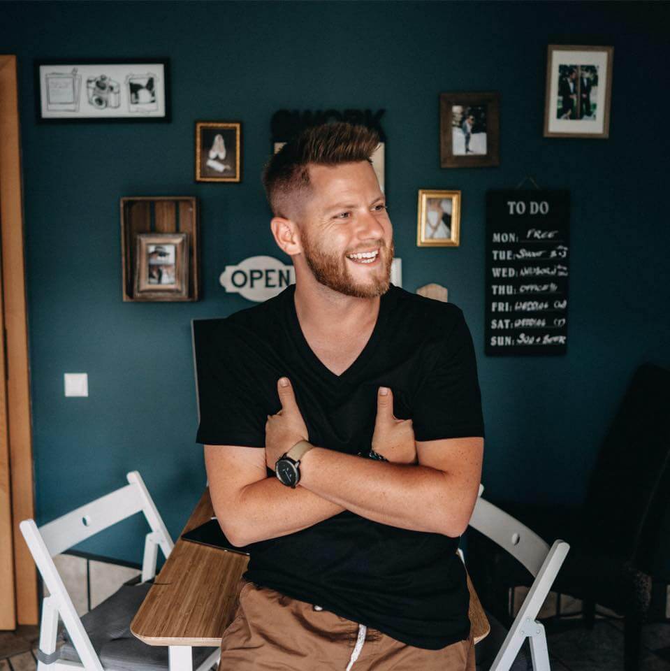 Ein lächelnder Mann mit kurzen Haaren und Bart, der ein schwarzes T-Shirt und eine braune Hose trägt, sitzt mit verschränkten Armen an einem Tisch in einem gemütlichen Raum, der mit gerahmten Fotos und einer To-Do-Liste an einer dunkelgrauen Wand dekoriert ist.