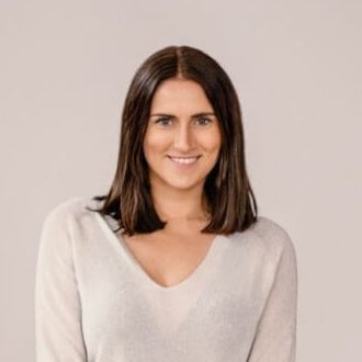 Eine Frau mit glattem braunem Haar, die ein hellgraues, langärmeliges Oberteil, weit geschnittene Jeans und Turnschuhe trägt, steht auf einem Bein und lächelt in die Kamera vor einem schlichten, hellen Hintergrund.