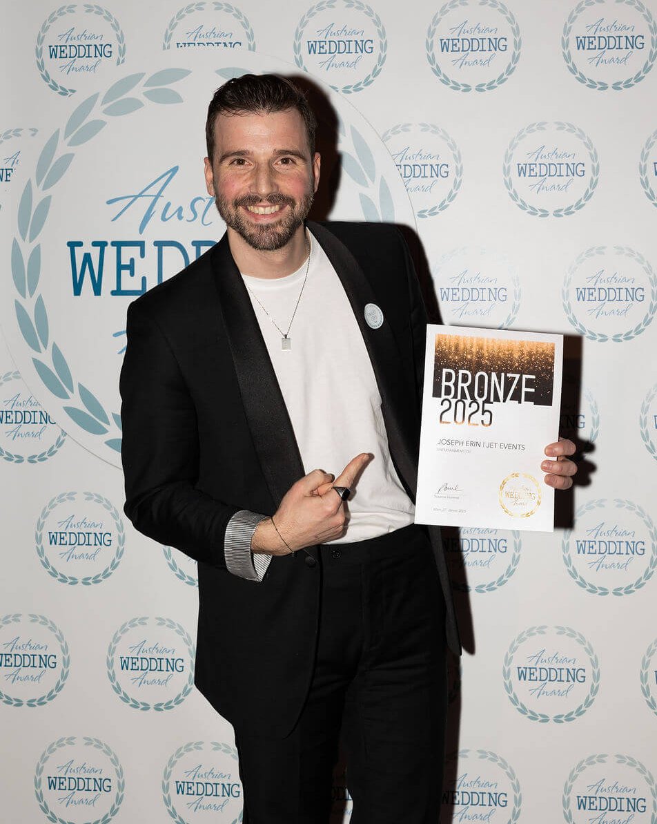Ein Mann in schwarzem Anzug und weißem Hemd lächelt und zeigt auf eine Urkunde mit der Aufschrift "Bronze 2025" vor einem Hintergrund mit sich wiederholenden "Australian Wedding Awards"-Logos.