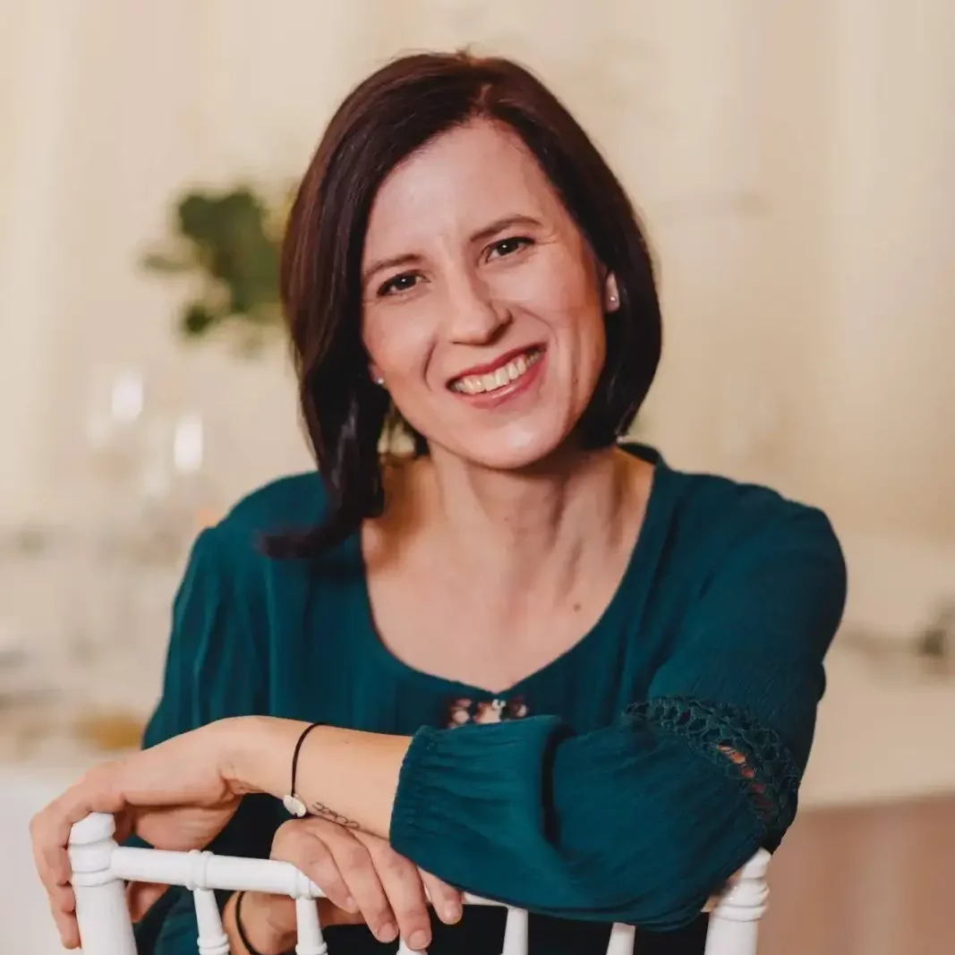 Eine Frau mit kurzen dunklen Haaren, die eine blaugrüne Bluse trägt, lächelt, während sie sich an die Lehne eines weißen Holzstuhls lehnt. Der Hintergrund ist leicht unscharf und zeigt einen Tisch mit Glaswaren und grünem Laub.