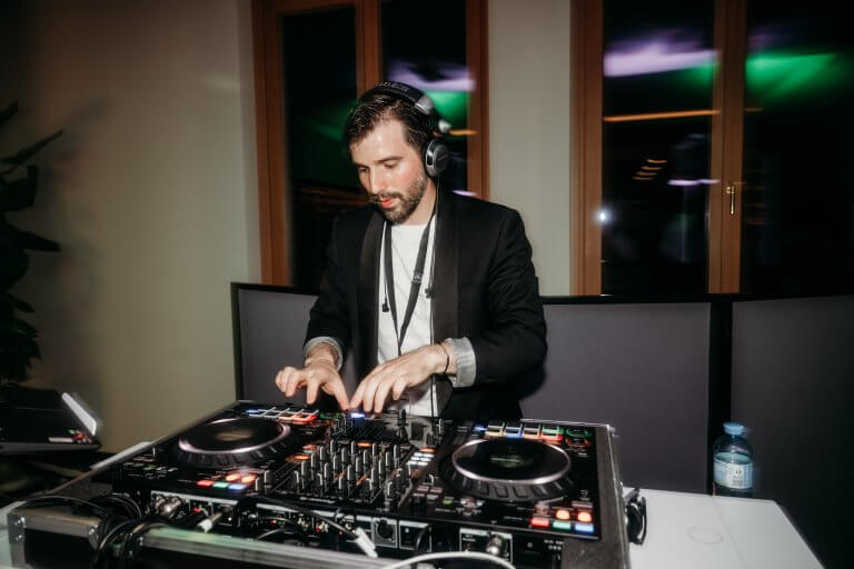 Ein DJ mit Kopfhörern und schwarzem Blazer mischt Musik an einem professionellen DJ-Controller bei einer Indoor-Veranstaltung, wobei sich bunte Lichter in den Fenstern hinter ihm spiegeln. Eine Flasche Wasser steht auf dem Tisch.
