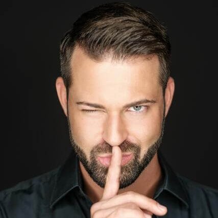 Ein Mann mit kurzen Haaren und Bart zwinkert, während er seinen Zeigefinger an die Lippen hält, um "pst" zu sagen. Er trägt ein dunkles Hemd mit Knöpfen vor einem schwarzen Hintergrund.
