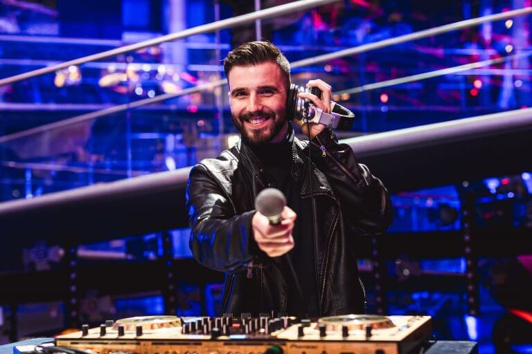 Ein lächelnder DJ in einer schwarzen Lederjacke steht hinter einem Mischpult, hält mit einer Hand ein Mikrofon in die Kamera und mit der anderen einen Kopfhörer ans Ohr, in einer lebhaften, bunten Clubumgebung.