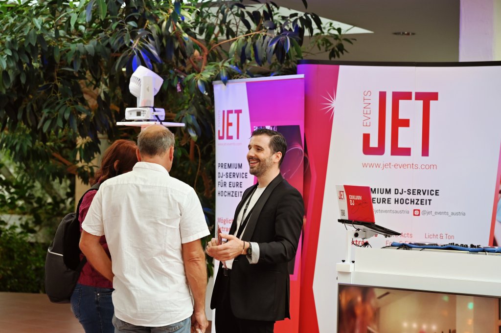 Ein lächelnder Mann im Anzug unterhält sich mit zwei Besuchern am Stand von JET Events auf der Hochzeitsmesse in der Eventpyramide Vösendorf, wo Premium-DJ-Services für Hochzeiten vorgestellt werden. Werbebanner, ein Laptop und Equipment sind im Hintergrund zu sehen.