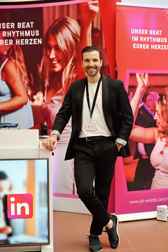 Ein Mann im Anzug posiert und lächelt an einem Veranstaltungsstand auf Hochzeitsmesse in der Eventpyramide Vösendorf mit rosa Bannern, die tanzende Menschen auf der hochzeitsmesse klagenfurt zeigen. Auf den Bannern hinter ihm steht "Unser Beat im Rhythmus eurer Herzen".