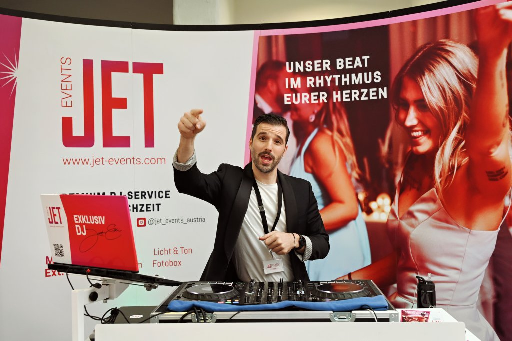 Ein DJ im Anzug steht an einem Mischpult und zeigt nach vorne, hinter ihm ein Laptop und Veranstaltungsbanner. Im Hintergrund sieht man lächelnde, tanzende Menschen und einen fetten Text, der für JET EVENTS auf der hochzeitsmesse Klagenfurt wirbt.
