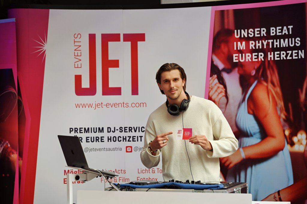 Ein Mann steht hinter einem DJ-Equipment, hält eine Karte in der Hand und lächelt. Hinter ihm wirbt ein JET Events-Banner für Premium-DJ-Services für Hochzeiten auf der hochzeitsmesse Klagenfurt. Ein rosa Plakat zeigt ein tanzendes Paar mit dem Text "Unser Beat im Rhythmus eurer Herzen".