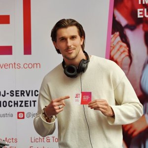 Ein Mann steht hinter einem DJ-Equipment, hält eine Karte in der Hand und lächelt. Hinter ihm wirbt ein JET Events-Banner für Premium-DJ-Services für Hochzeiten auf der hochzeitsmesse Klagenfurt. Ein rosa Plakat zeigt ein tanzendes Paar mit dem Text "Unser Beat im Rhythmus eurer Herzen".