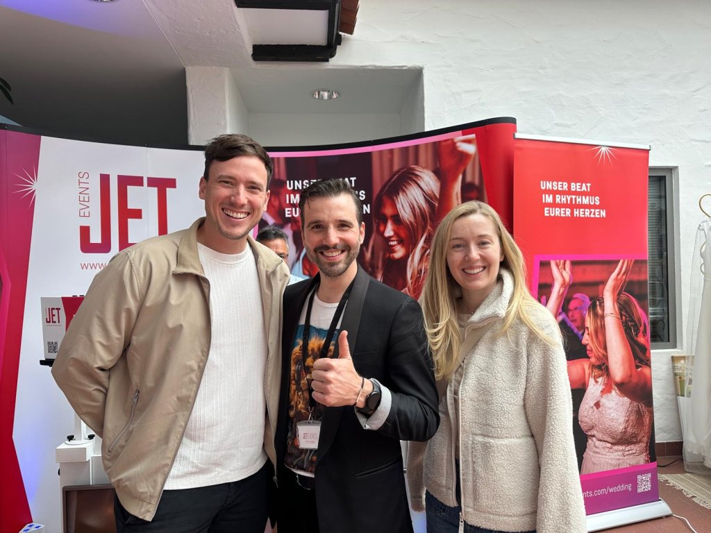 Drei lächelnde Menschen stehen bei einer Indoor-Veranstaltung zusammen, wahrscheinlich die hochzeitsmesse Klagenfurt; der Mann in der Mitte zeigt den Daumen hoch. Hinter ihnen sind Banner mit Partybildern und deutschem Text zu sehen. Die Umgebung ist hell und leger.