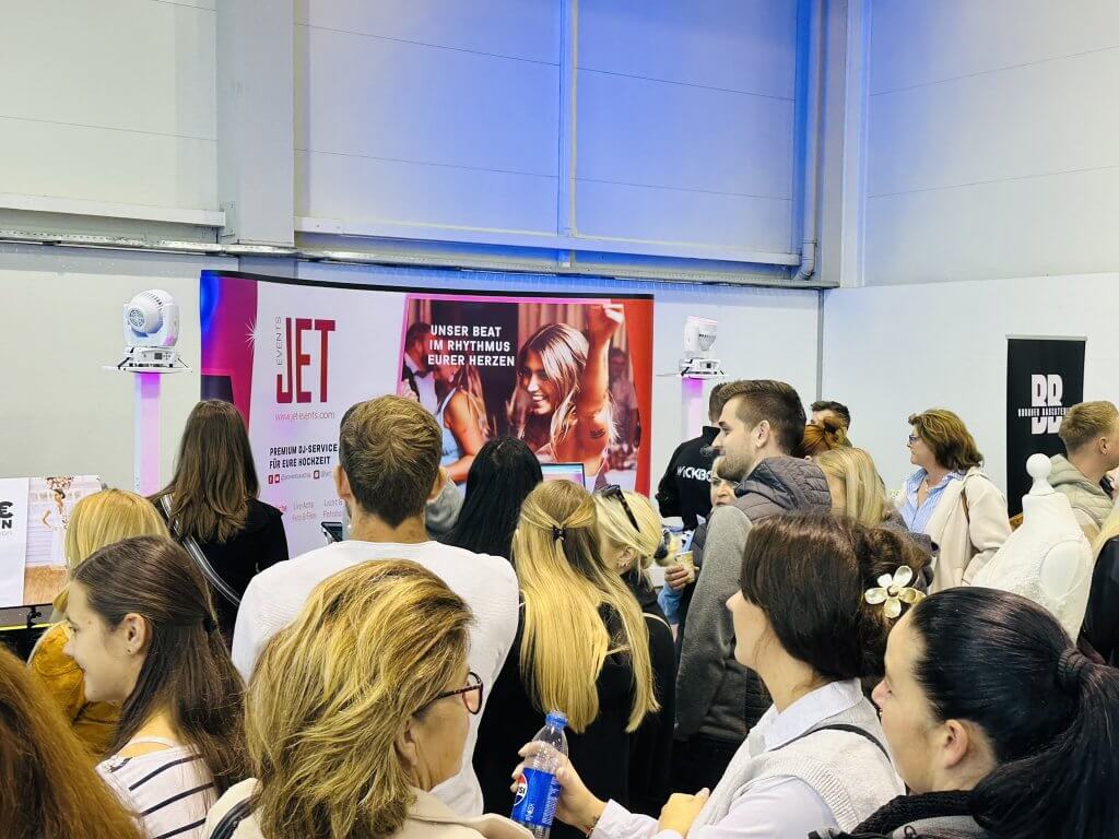 Eine Menschenmenge steht drinnen auf einer Veranstaltung vor einem Stand mit einem roten Schild mit der Aufschrift "JET" und einem Bild von tanzenden Menschen. Die Teilnehmer sind beschäftigt und hören einem Redner am Stand zu.