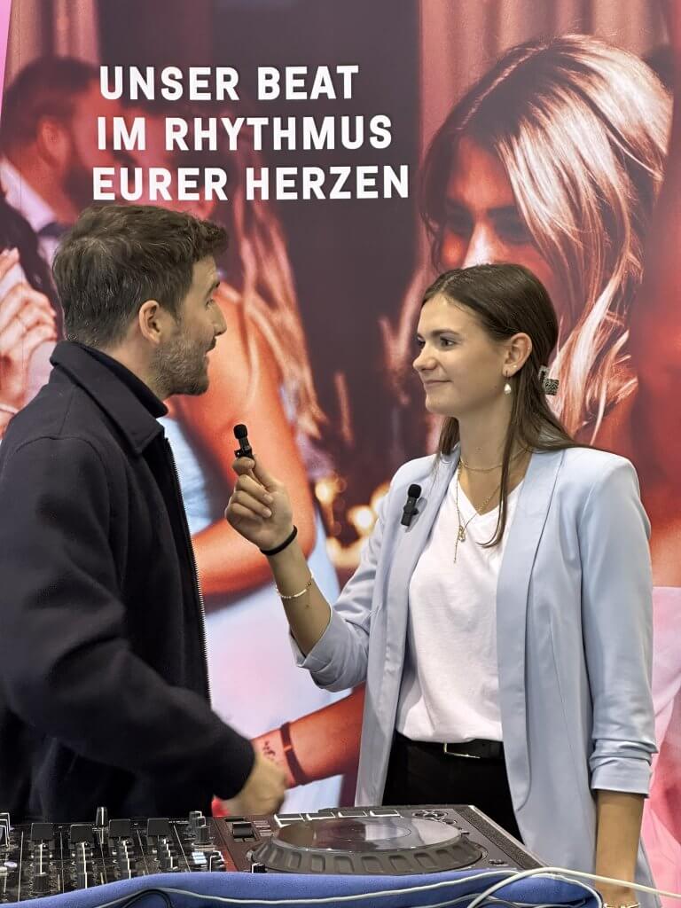 Eine Frau in einem hellblauen Blazer hält einem Mann in einer schwarzen Jacke ein Mikrofon entgegen. Sie stehen vor einer DJ-Anlage und einem Banner mit der Aufschrift "UNSER BEAT IM RHYTHMUS EURER HERZEN". Im Hintergrund sind unscharfe Menschen zu sehen.