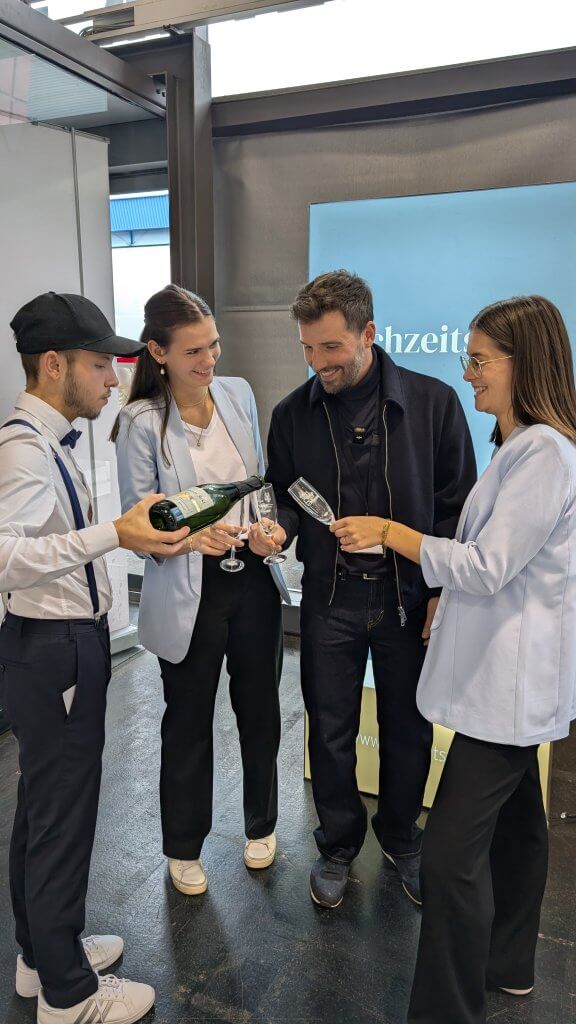 Vier Personen stehen in einem Innenraum zusammen und schenken lächelnd Sekt in Gläser ein. Zwei Frauen und zwei Männer sind elegant gekleidet und scheinen zu feiern oder ein geselliges Beisammensein zu genießen. Im Hintergrund ist eine blaue Fahne zu sehen.
