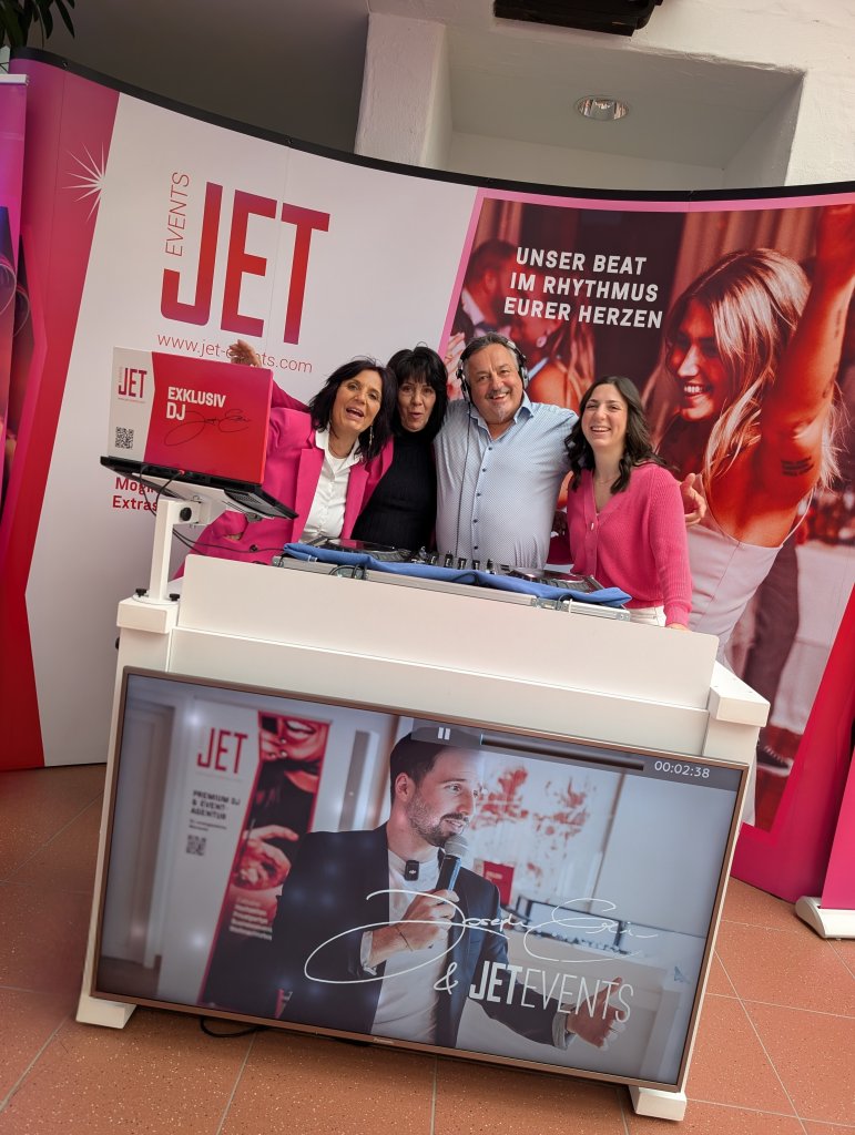 Vier lächelnde Erwachsene stehen gemeinsam hinter einem DJ-Pult mit "JET"-Branding auf der hochzeitsmesse klagenfurt. Ein großes Display zeigt tanzende Menschen und deutsche Slogans, die eine festliche und werbliche Atmosphäre für JET EVENTS schaffen.