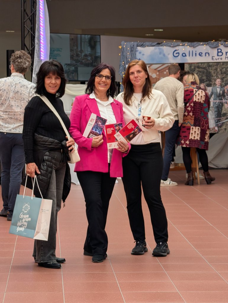 Drei Frauen stehen in einem Innenraum der hochzeitsmesse klagenfurt auf einem Kachelboden und lächeln in die Kamera. Zwei halten Prospekte in der Hand, eine trägt eine Einkaufstasche. Im Hintergrund sind weitere Personen und ein Stand zu sehen.