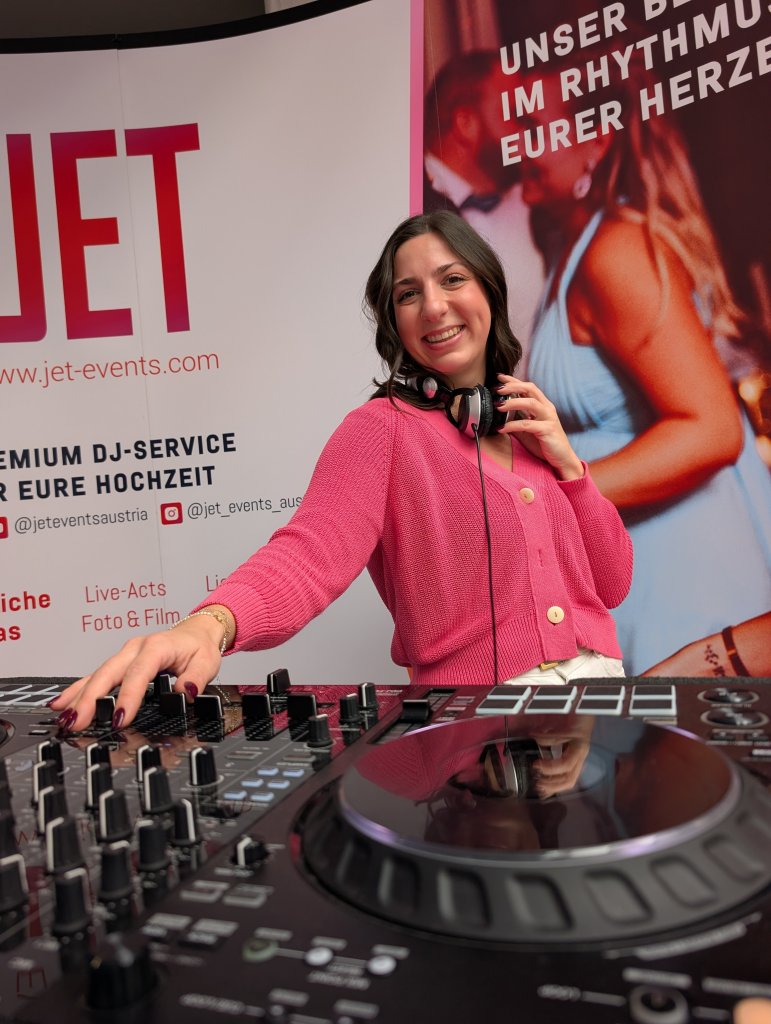 Eine lächelnde Frau in einem rosa Outfit steht hinter einem DJ-Controller und trägt Kopfhörer um den Hals. Sie legt an einem Veranstaltungsstand auf, der mit Bannern für einen Premium-DJ-Service auf der Hochzeitsmesse Klagenfurt wirbt.