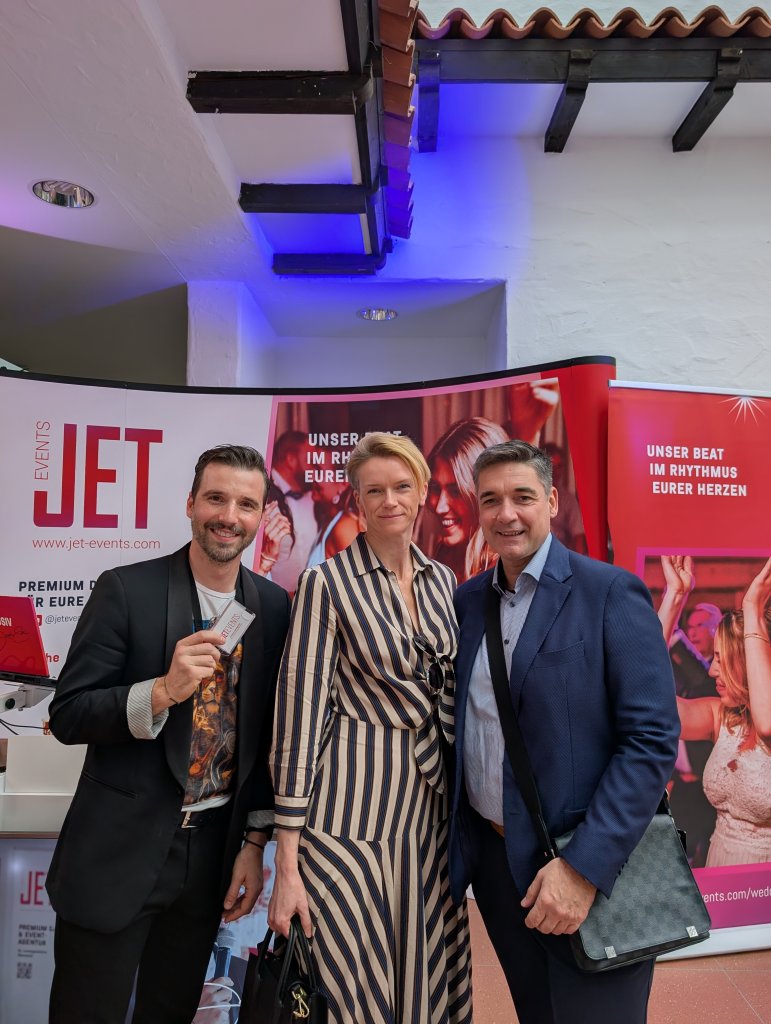 Drei formal gekleidete Personen stehen und lächeln bei einer Indoor-Veranstaltung. Sie posieren vor einem rot-weißen "JET Events"-Display auf der hochzeitsmesse klagenfurt, mit Partybildern und deutschem Text im Hintergrund.
