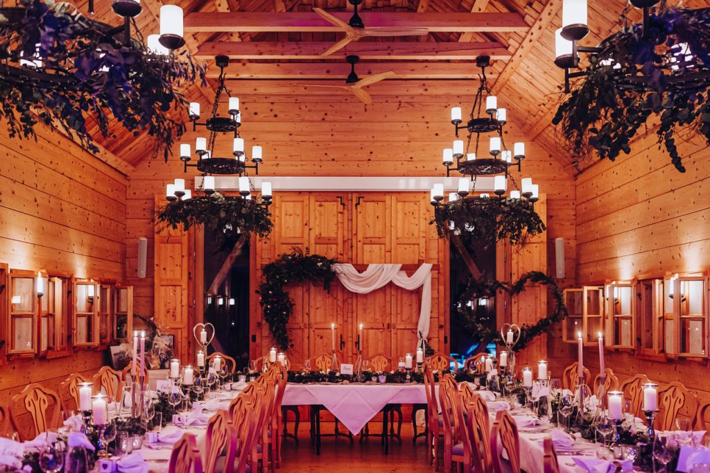 Ein warm beleuchteter hölzerner Festsaal, dekoriert mit Kronleuchtern, Grünzeug und Kerzen, eingerichtet für eine Winterhochzeit in Österreich - perfekt für einen bezaubernden Abend mit einem DJ Berghochzeit und zwei langen Tischen unter einem schönen drapierten Bogen.