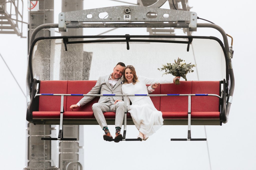 Eine Braut und ein Bräutigam in Hochzeitskleidung sitzen eng beieinander auf einem roten Skilift, lächelnd und mit einem Blumenstrauß in der Hand, wobei ein nebliger, grauer Hintergrund und die Liftmaschinen zu sehen sind.
