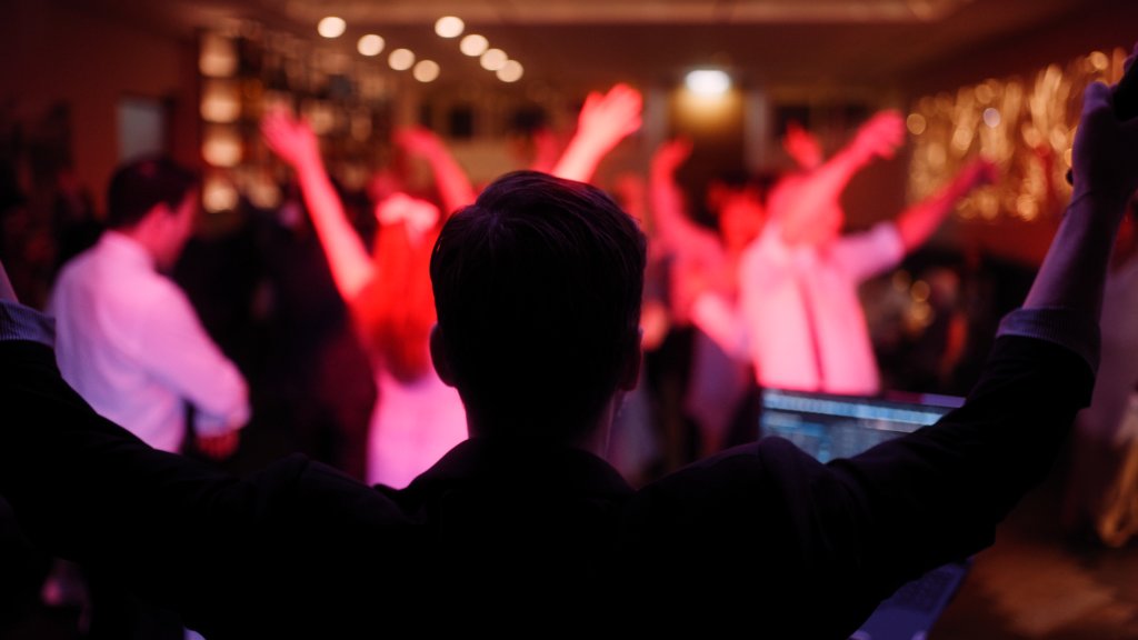 Eine Person mit erhobenen Armen steht vor einer tanzenden Menschenmenge in einem schwach beleuchteten Raum mit bunten Lichtern, möglicherweise auf einer Party oder einer Veranstaltung. Die Szene strahlt eine lebhafte, energiegeladene Atmosphäre aus.