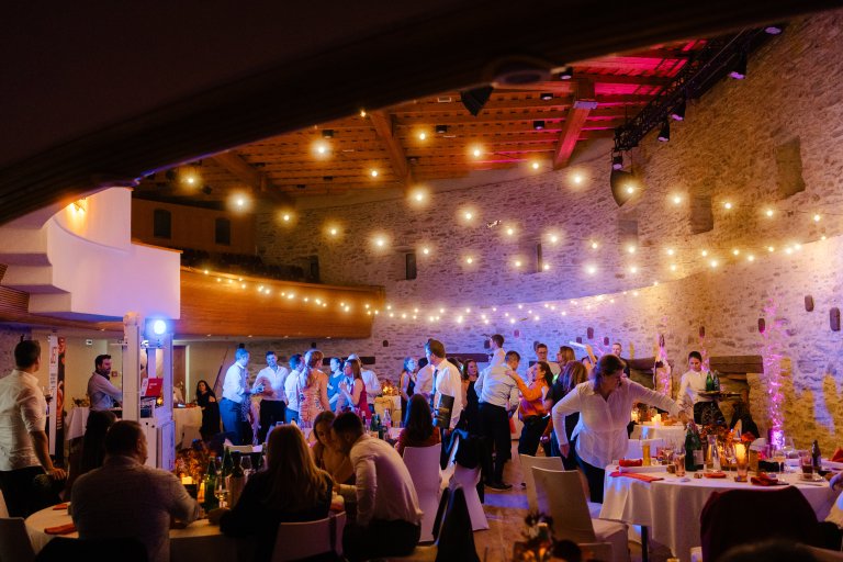 Eine lebhafte Hochzeitsfeier in einem Saal, bei der die Gäste unter warmer Lichterkette in einem rustikalen Saal aus Stein speisen und tanzen. Die Tische sind geschmückt, und alle genießen das Fest.