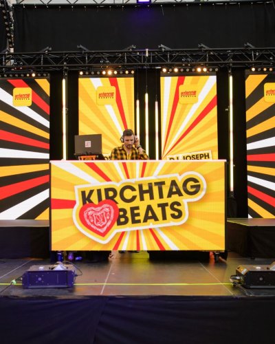 Ein DJ legt auf einer hell erleuchteten Bühne auf, auf der ein "KIRCHTAG BEATS"-Banner, bunte Strahlen im Hintergrund und eine Tonanlage zu sehen sind. Links steht ein Wachmann, rechts ist eine Treppe zu sehen.