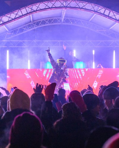 Eine winterlich gekleidete Menschenmenge steht nachts vor einer hell erleuchteten Bühne, auf der ein professioneller DJ mit einem Helm einen Arm hebt. Bunte Lichter und Nebel schaffen eine lebendige, energiegeladene Atmosphäre, die perfekt für jede Veranstaltung ist, in der ein DJ auftritt.