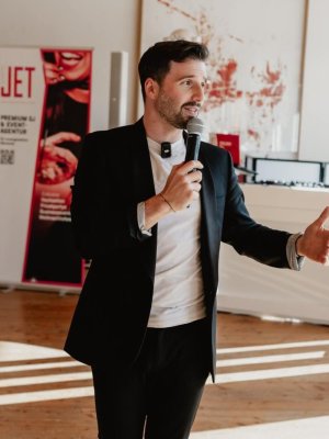 Ein Mann im schwarzen Anzug hält ein Mikrofon und gestikuliert, während er in einem hellen Raum mit Holzböden, moderner Kunst an den Wänden und einer stilvollen Atmosphäre spricht, die perfekt für Musik und DJs für Feiern in Österreich ist.