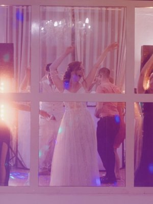 Eine Gruppe von Menschen, darunter eine Braut in einem weißen Kleid, tanzt fröhlich zusammen im Innenbereich hinter einem großen Fenster, das von warmen, bunten Lichtern beleuchtet wird.