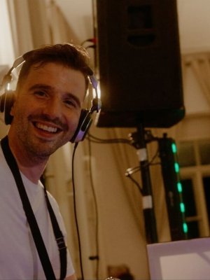 Hochzeits DJ Joseph Erin mit Kopfhörern und weißem Hemd lächelt und streckt die Hand in die Kamera. Sie befindet sich in einem Raum mit Vorhängen und steht neben einer Audioanlage und einem Lautsprecher.