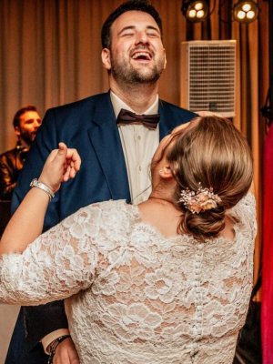 Ein Paar tanzt fröhlich auf einer Hochzeitsfeier. Der Bräutigam in einem marineblauen Anzug und die Braut in einem Spitzenbrautkleid lachen zusammen. Im Hintergrund sind ein DJ-Pult und rosa Banner mit Text auf Deutsch zu sehen.