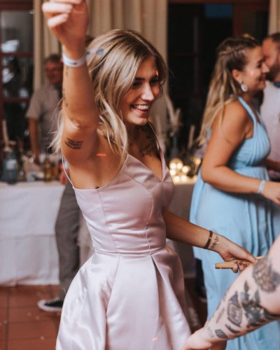 Eine Frau mit blonden Haaren und Tattoos, die ein rosa Kleid trägt, tanzt fröhlich auf einer lebhaften Indoor-Party, umgeben von anderen Gästen.
