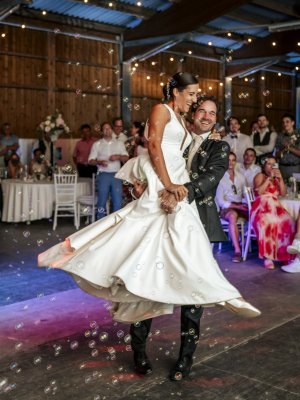 Ein Bräutigam hebt und dreht seine lächelnde Braut auf der Tanzfläche während ihres Hochzeitsempfangs in einer rustikalen Scheune, während ein Hochzeits-DJ die Luft mit Musik füllt und die Gäste, die an dekorierten Tischen sitzen, inmitten schwebender Seifenblasen applaudieren.