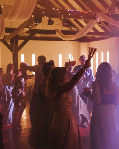 Eine Gruppe von Menschen tanzt in einem warm beleuchteten Raum mit drapiertem Stoff und Lichterketten über dem Kopf. Silhouetten und weiches Licht schaffen eine festliche, feierliche Atmosphäre.