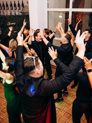 Eine Gruppe von Menschen in formeller Kleidung tanzt begeistert auf einer Party in einem gut beleuchteten Veranstaltungsort. Sie haben die Arme erhoben und wirken fröhlich. Der Raum hat einen Holzboden, weiße Wände und im Hintergrund sind Weinflaschen ausgestellt.